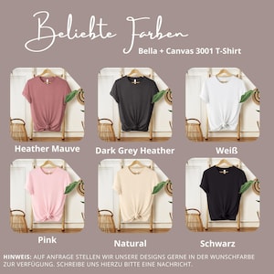 May include: A display of six Bella + Canvas 3001 T-shirts in various colors, including heather mauve, dark grey heather, white, pink, natural, and black. Each shirt is knotted at the waist and displayed on a wooden hanger.
