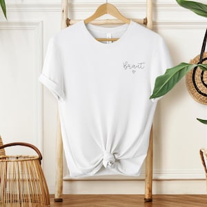 May include: A white t-shirt with the word "Bride" in script and a small heart. The shirt is knotted at the waist and hangs on a wooden hanger. The shirt is displayed on a wooden ladder.