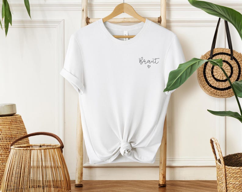 May include: A white t-shirt with the word "Bride" in script and a small heart. The shirt is knotted at the waist and hangs on a wooden hanger. The shirt is displayed on a wooden ladder.