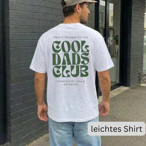 May include: A white short-sleeved t-shirt featuring dark green text on the back that reads PROUD MEMBER OF THE COOL DADS CLUB and POWERED BY LOVE & ESPRESSO in a stylized font. The shirt is paired with light blue denim jeans and a tan baseball cap.