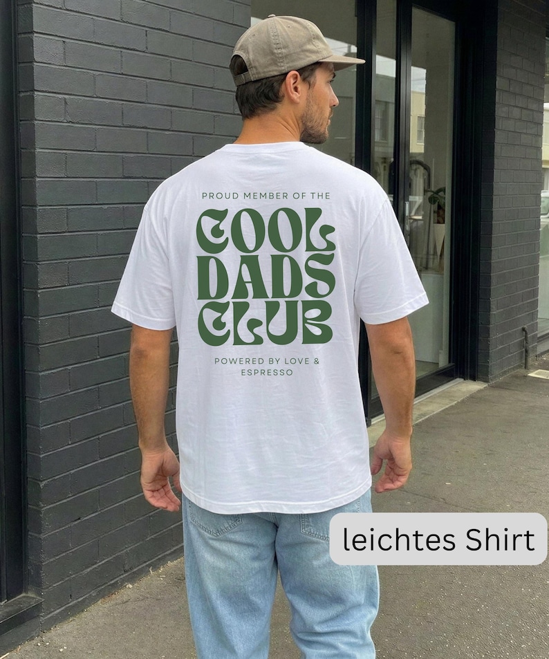 May include: A white short-sleeved t-shirt featuring dark green text on the back that reads PROUD MEMBER OF THE COOL DADS CLUB and POWERED BY LOVE & ESPRESSO in a stylized font. The shirt is paired with light blue denim jeans and a tan baseball cap.