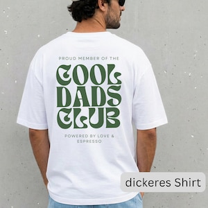 May include: A white short-sleeved t-shirt featuring dark green text on the back that reads PROUD MEMBER OF THE COOL DADS CLUB and POWERED BY LOVE & ESPRESSO in a stylized font. The shirt is paired with light blue denim jeans.