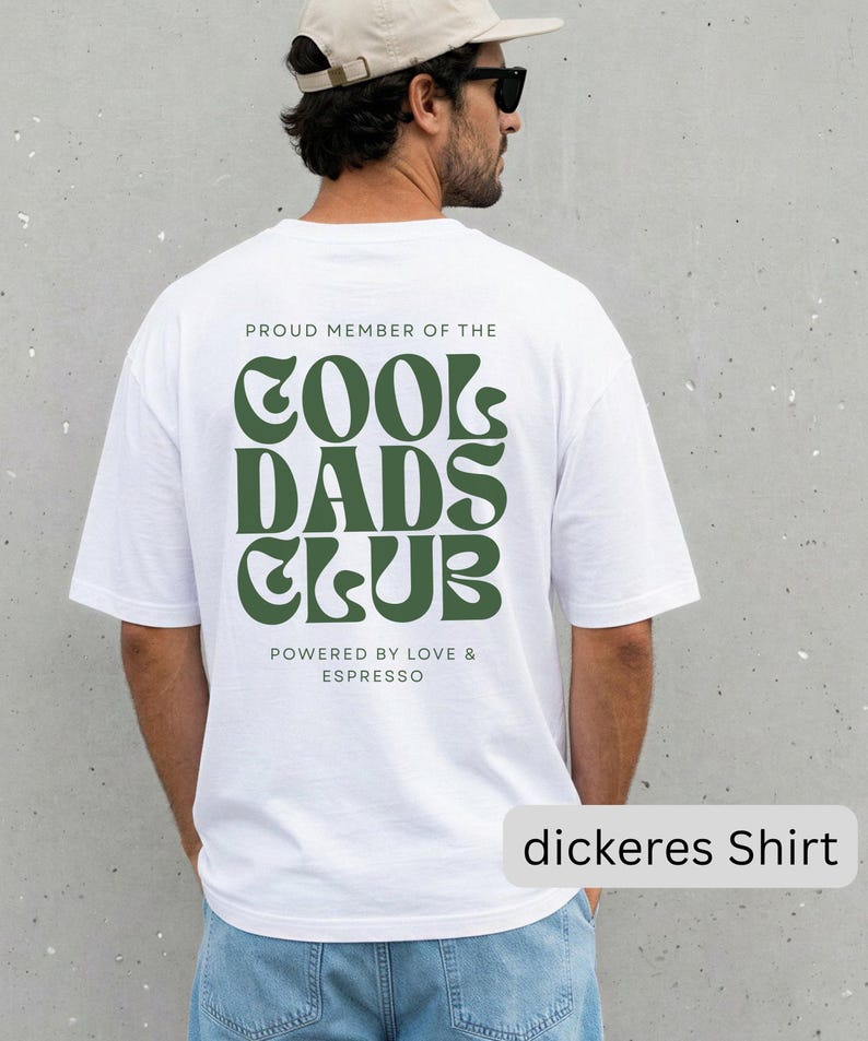 May include: A white short-sleeved t-shirt featuring dark green text on the back that reads PROUD MEMBER OF THE COOL DADS CLUB and POWERED BY LOVE & ESPRESSO in a stylized font. The shirt is paired with light blue denim jeans.