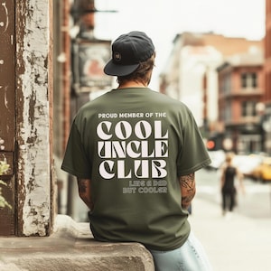Könnte beinhalten: Ein olivgrünes T-Shirt mit dem weißen Text "PROUD MEMBER OF THE COOL UNCLE CLUB LIKE A DAD BUT COOLER". Eine Person mit einer schwarzen Baseballkappe ist von hinten zu sehen, die sich im Freien aufhält.