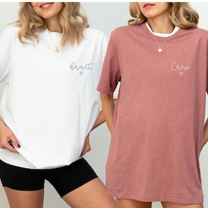 May include: Two women wearing matching t-shirts. One shirt is white with the word "Braut" and a heart, the other is heather mauve with "Crew" and a heart. Both shirts are short-sleeved.