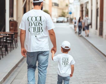 May include: A father and child walk down a city street, both wearing white t-shirts and baseball caps. The father's shirt reads "COOL DADS CLUB" and the child's shirt says "COOL KIDS CLUB". Both are wearing blue jeans and white sneakers.