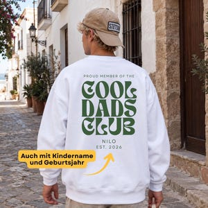 Könnte beinhalten: Ein weißes Sweatshirt mit Rundhalsausschnitt und dunkelgrünem Text auf der Rückseite, der PROUD MEMBER OF THE COOL DADS CLUB lautet, mit dem Text NILO EST. 2026 darunter. Das Sweatshirt wird mit einer beigen Baseballkappe und einer hellen Hose getragen.