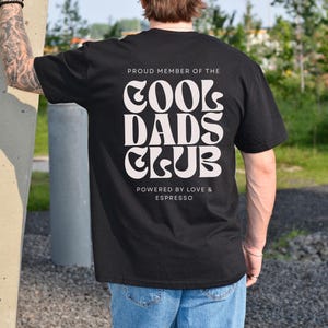 May include: A black short-sleeved t-shirt featuring white text on the back that reads PROUD MEMBER OF THE COOL DADS CLUB, with the additional text below stating POWERED BY LOVE & ESPRESSO in a clean, sans-serif font style.