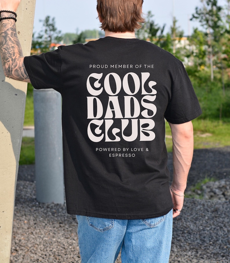 May include: A black short-sleeved t-shirt featuring white text on the back that reads PROUD MEMBER OF THE COOL DADS CLUB, with the additional text below stating POWERED BY LOVE & ESPRESSO in a clean, sans-serif font style.