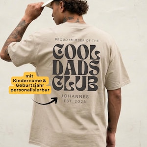 Könnte beinhalten: Ein beiges T-Shirt mit kurzen Ärmeln und dunkelbraunem Text auf der Rückseite, der PROUD MEMBER OF THE COOL DADS CLUB lautet, sowie dem Namen JOHANNES und dem Text EST. 2026, die unter dem Hauptmotiv in einer serifenlosen Schrift gedruckt sind.