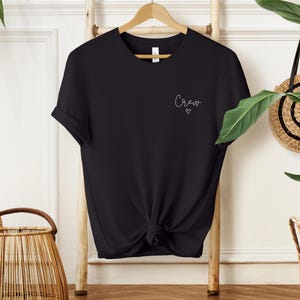 May include: Black t-shirt with rolled sleeves, knotted at the waist, hanging on a wooden hanger. The word "Crew" is written in cursive on the upper left chest, with a small heart below it. The shirt is displayed against a white background.