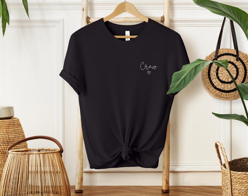 May include: Black t-shirt with rolled sleeves, knotted at the waist, hanging on a wooden hanger. The word "Crew" is written in cursive on the upper left chest, with a small heart below it. The shirt is displayed against a white background.