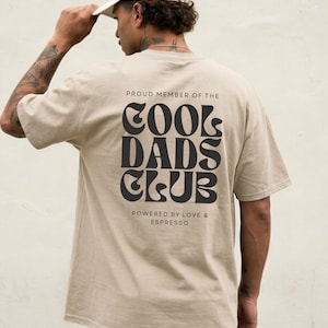 Könnte beinhalten: Ein beiges Kurzarm-T-Shirt mit schwarzem Text auf der Rückseite, der PROUD MEMBER OF THE COOL DADS CLUB lautet, sowie dem zusätzlichen Text POWERED BY LOVE & ESPRESSO in einer kleineren Schriftart unter dem Hauptmotiv.