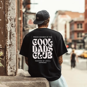 May include: A black short-sleeve t-shirt featuring white text on the back that reads PROUD MEMBER OF THE COOL DADS CLUB POWERED BY LOVE & ESPRESSO in a bold, wavy font. The shirt is worn with a dark grey baseball cap, viewed from the back.