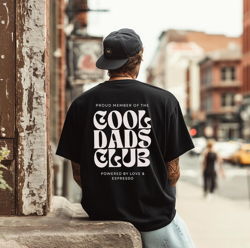 May include: A black short-sleeve t-shirt featuring white text on the back that reads PROUD MEMBER OF THE COOL DADS CLUB POWERED BY LOVE & ESPRESSO in a bold, wavy font. The shirt is worn with a dark grey baseball cap, viewed from the back.