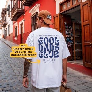 Könnte beinhalten: Ein weißes T-Shirt mit kurzen Ärmeln und dunkelblauem Text auf der Rückseite, der PROUD MEMBER OF THE COOL DADS CLUB lautet, gefolgt vom Namen JOHANNES und dem Text EST. 2026. Das T-Shirt wird mit einer beigen Baseballkappe mit weißem Aufnäher getragen.