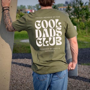 May include: An olive green short-sleeve t-shirt featuring white text on the back that reads PROUD MEMBER OF THE COOL DADS CLUB and POWERED BY LOVE & ESPRESSO in a bold, wavy, retro-inspired font style with a clean, sans-serif typeface below.