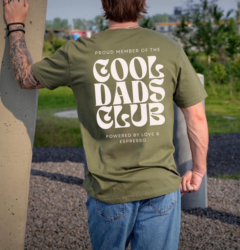 May include: An olive green short-sleeve t-shirt featuring white text on the back that reads PROUD MEMBER OF THE COOL DADS CLUB and POWERED BY LOVE & ESPRESSO in a bold, wavy, retro-inspired font style with a clean, sans-serif typeface below.