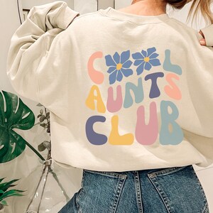 Könnte beinhalten: Cremefarbenes Sweatshirt mit dem Text "COOL AUNTS CLUB" in farbenfrohen Buchstaben im Retro-Stil. Das Design beinhaltet zwei blaue Blumengrafiken. Das Sweatshirt wird mit einer blauen Jeans kombiniert.