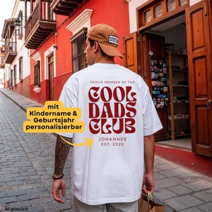 Könnte beinhalten: Ein weißes T-Shirt mit kurzen Ärmeln und dunkelrotem Text auf der Rückseite: PROUD MEMBER OF THE COOL DADS CLUB, JOHANNES, EST. 2026. Das T-Shirt wird mit einer beigen Baseballkappe getragen. Ein gelbes Grafik-Overlay enthält deutschen Text.
