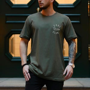 Könnte beinhalten: Olivgrünes T-Shirt mit dem weißen Schriftzug "Cool Uncles Club" auf der linken Brust. Das Shirt hat kurze Ärmel und einen Rundhalsausschnitt. Die Person trägt eine schwarze Hose und eine Uhr.