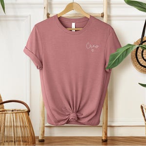 May include: A dusty rose t-shirt with rolled sleeves is displayed on a wooden hanger. The word "Crew" is written in cursive with a small heart underneath. The shirt is knotted at the bottom.