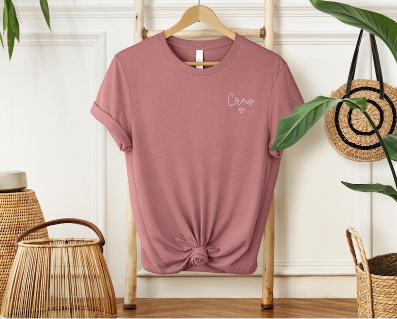 May include: A dusty rose t-shirt with rolled sleeves is displayed on a wooden hanger. The word "Crew" is written in cursive with a small heart underneath. The shirt is knotted at the bottom.