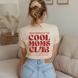 Könnte beinhalten: Ein cremefarbenes Kurzarm-T-Shirt mit rotem Text auf der Rückseite, der PROUD MEMBER OF THE COOL MOMS CLUB und RUNNING ON CUDDLES AND FUN lautet. Das T-Shirt wird mit einer hellblauen Jeans kombiniert, die einen beigen Lederaufnäher hinten hat.