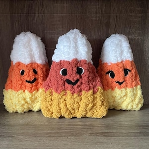 Puede incluir: Tres decoraciones de felpa de maíz de caramelo. Cada una tiene una parte superior blanca, un centro coral y una parte inferior amarilla. Tienen caras sonrientes negras. Los peluches están hechos de un material suave y texturizado.