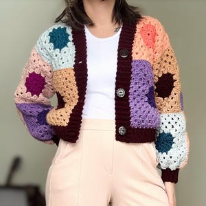 Puede incluir: Un cárdigan de ganchillo colorido con un diseño de patchwork. El cárdigan tiene un ribete y botones marrones. El cárdigan se lleva sobre un top blanco sin mangas y pantalones rosas.