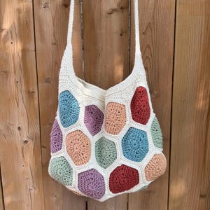May include: A handmade crochet bag with a cream-colored base and a colorful honeycomb pattern. The bag features a variety of pastel colors, including blue, pink, green, and purple. It has a long, cream-colored strap for over-the-shoulder wear.