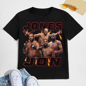 Può includere: Maglietta nera con una grafica di quattro uomini in posizione di boxe. Il testo "JONES JON" è stampato in rosso e nero sulla maglietta.