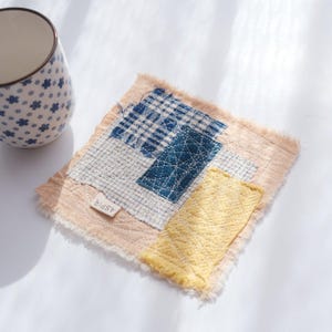 May include: A square, patchwork coaster with frayed edges. The coaster features patches of cream, blue plaid, and yellow fabric, stitched together. A small tag with the text "ASPR" is attached. A blue and white floral patterned mug is in the background.