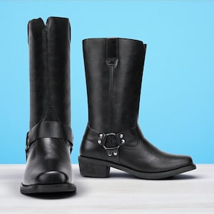 May include: A pair of black leather motorcycle boots. The boots have a square toe, a mid-height heel, and a strap with a metal buckle around the ankle. The shaft of the boots reaches mid-calf. The background is a gradient of blue.