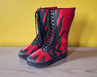 May include: A pair of tall, red and black boots with a flame design. The boots have a lace-up closure and a flat sole. The red sections are outlined with black, creating a bold contrast. The boots are made of a smooth material and appear to be new.