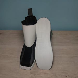 Peut inclure: Une paire de bottes noires, blanches et grises. Une botte est droite, l'autre est couch&eacute;e sur le c&ocirc;t&eacute;, r&eacute;v&eacute;lant la semelle blanche. Les bottes ont un design moderne avec une silhouette montante et une tirette &agrave; l'arri&egrave;re.