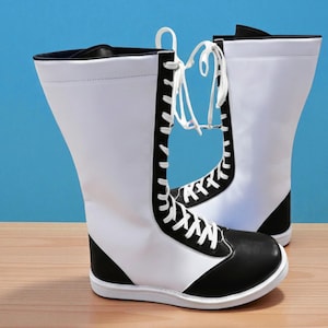 May include: A pair of white and black boots with a unique design. The boots have a white upper with black accents on the toe and heel. White laces run up the front. The boots are on a wooden surface against a blue background.