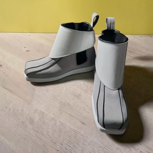 May include: A pair of gray ankle boots with white accents. The boots have a unique design with a white fold-over cuff and black vertical stripes on the front. The soles are white, and the boots have a pull tab at the back.