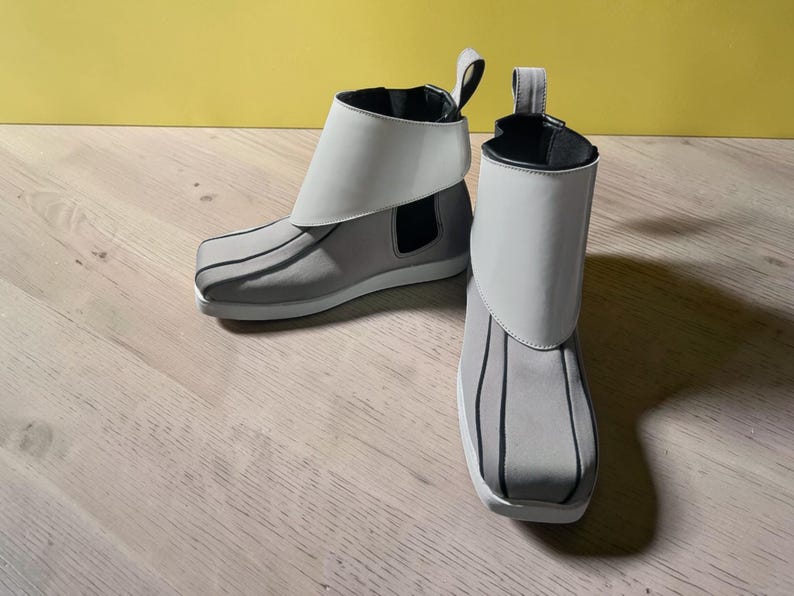 May include: A pair of gray ankle boots with white accents. The boots have a unique design with a white fold-over cuff and black vertical stripes on the front. The soles are white, and the boots have a pull tab at the back.