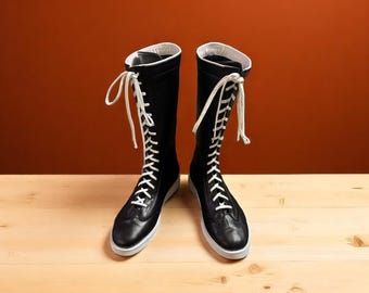 May include: A pair of black knee-high boots with white laces and white trim. The boots are made of leather and have a white sole. The background is a solid orange-brown color, and the boots are on a light wood surface.