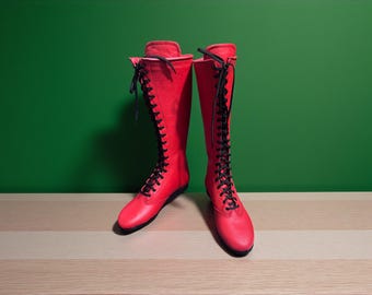 May include: A pair of knee-high red boots with black laces. The boots are made of a smooth material and have a flat sole. The boots are standing on a light brown and white striped surface against a green background.