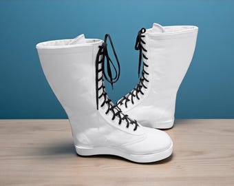 Pode incluir: Um par de botas brancas de cano alto com atacadores pretos. As botas são feitas de um material liso e têm uma biqueira arredondada. O fundo é azul liso e as botas estão sobre uma superfície de madeira clara.