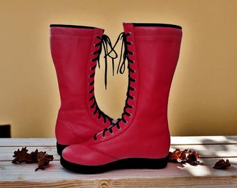 May include: A pair of vibrant red, knee-high boots with black laces and soles. The boots have a classic design, with a smooth, solid color and a lace-up front. The boots are displayed on a wooden surface.