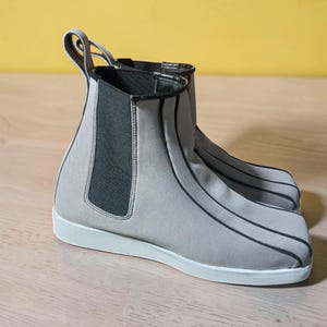 May include: A pair of gray Chelsea boots with black trim and white soles. The boots have a pull tab at the back and elastic side panels. The boots are made of a soft, flexible material.