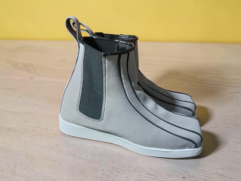 May include: A pair of gray Chelsea boots with black trim and white soles. The boots have a pull tab at the back and elastic side panels. The boots are made of a soft, flexible material.