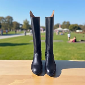 Puede incluir: Un par de botas de montar negras de cuero. Las botas son altas y tienen un diseño clásico. Se exhiben sobre una superficie de madera, con un parque y cielo azul al fondo. Las botas miden aproximadamente 40 cm de altura.