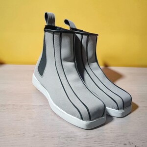 May include: A pair of gray ankle boots with black vertical stripes and white soles. The boots have a pull tab at the back and a black elastic panel on the side. The background is a solid yellow.