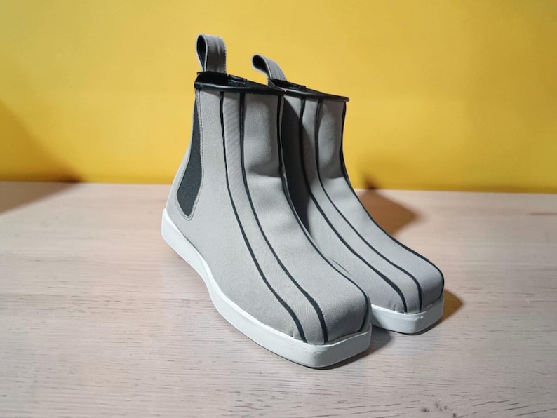 May include: A pair of gray ankle boots with black vertical stripes and white soles. The boots have a pull tab at the back and a black elastic panel on the side. The background is a solid yellow.