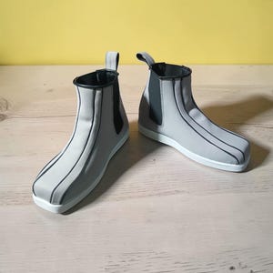 May include: A pair of gray ankle boots with black trim and accents. The boots have a pull tab at the back and a white sole. The boots are made of a smooth material and are designed with vertical black lines.