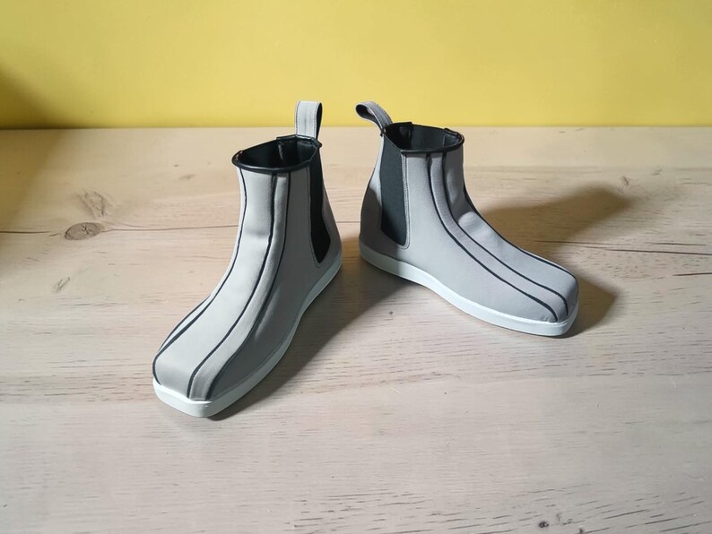 May include: A pair of gray ankle boots with black trim and accents. The boots have a pull tab at the back and a white sole. The boots are made of a smooth material and are designed with vertical black lines.